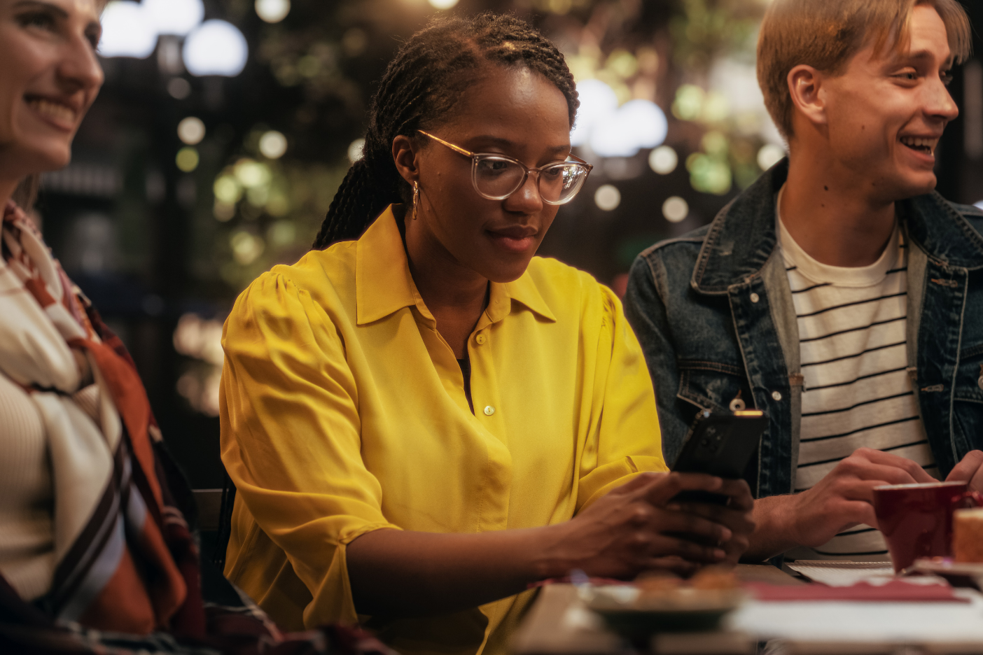Young Black Female Focused On Smartphone, Not Paying Attention to a Group of Friends Sitting in a Cafe Terrace Around Her. African Girl is Zoned Out, Entertained by Online Social Media on Her Device